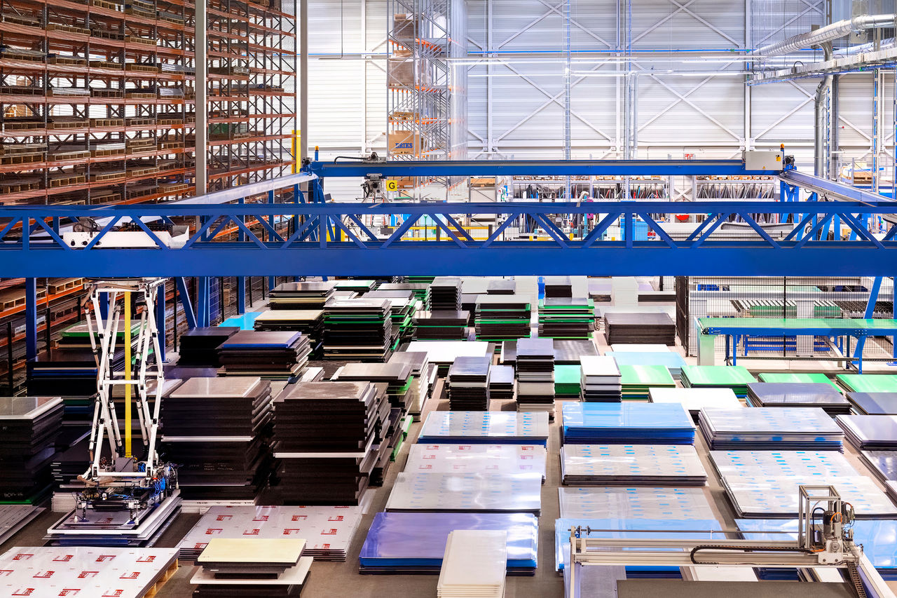 Een ruime opslagloods gevuld met netjes gestapelde plastic platen in verschillende kleuren en maten. Een blauwe bovenloopkraan is zichtbaar, met stellingen en industriële apparatuur op de achtergrond.