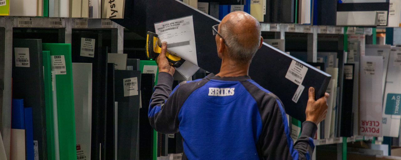 Een ERIKS-medewerker in een blauw uniform haalt een gelabelde plastic plaat van een schap, omringd door diverse monsters in verschillende kleuren en maten.