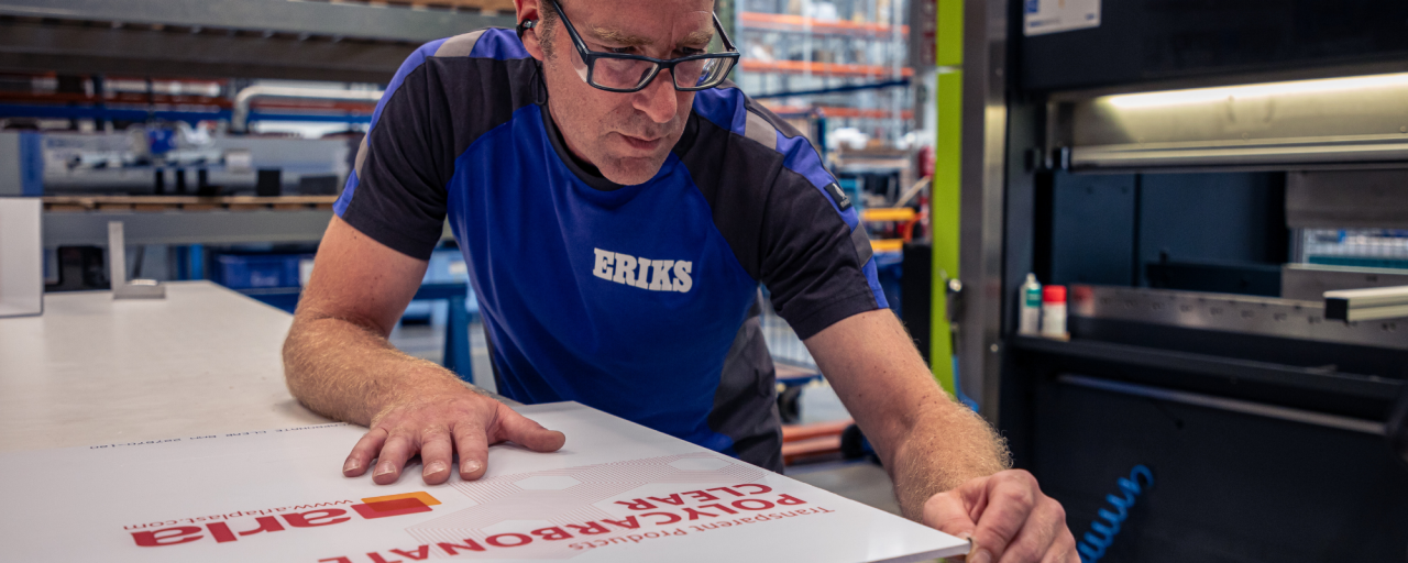 Een ERIKS-medewerker in een blauw uniform onderzoekt nauwkeurig een polycarbonaat plaat op een werkbank in een magazijnomgeving.