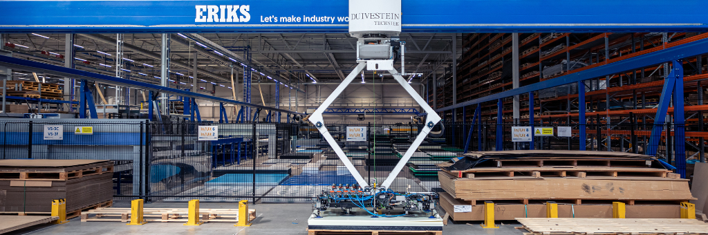 Een grote ERIKS-bovenloopkraan in een ruime opslagplaats, uitgerust met een robotisch heftmechanisme, omringd door netjes gestapelde pallets en stellingen.