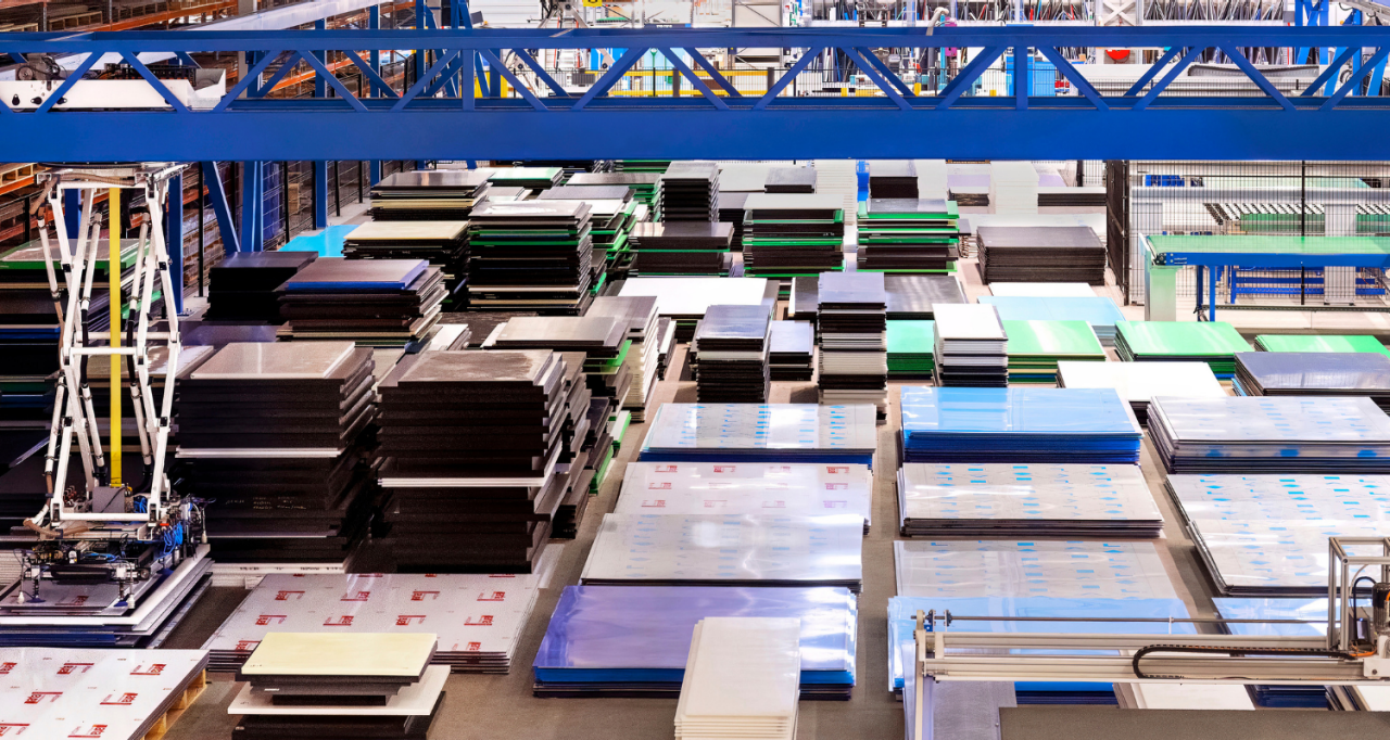 Een ruime opslagloods gevuld met netjes gestapelde plastic platen in verschillende kleuren en maten. Een blauwe bovenloopkraan is zichtbaar, met stellingen en industriële apparatuur op de achtergrond.
